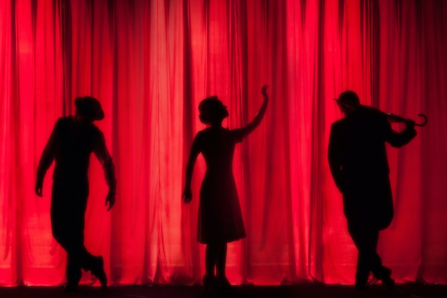 sihouette of a woman dancing with two male silhouettes on either side of her. they are behind a red, backlit stage curtain