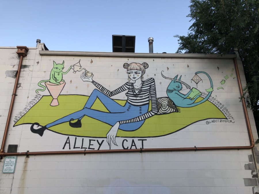 painting of woman sitting on green rug drinking coffee surrounded by cats, with the words Alley Cat written below