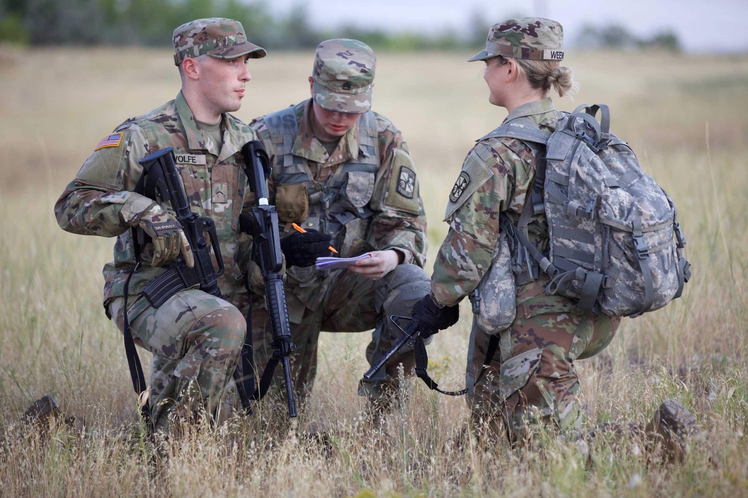 three people in military uniforms holding guns kneel in field
