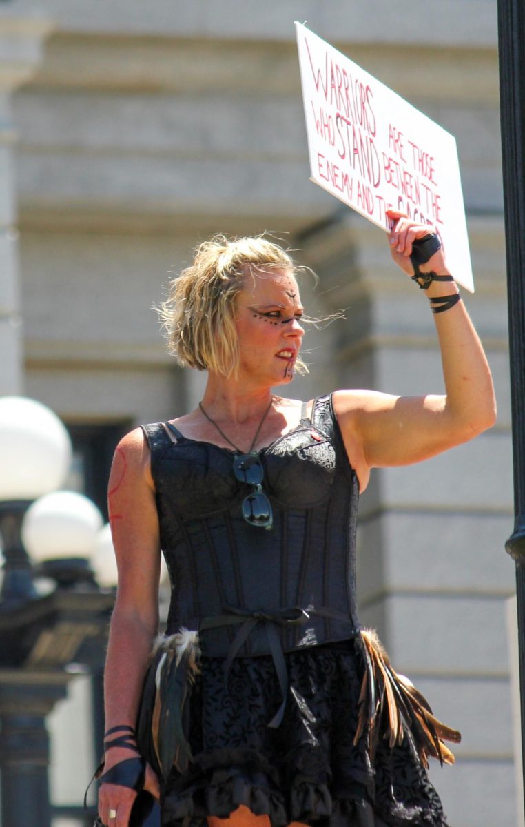 A protestor dressed as a warrior stands on the steps of the Denver Capital holding a sign that says, Warriors are those who stand between the enemies and the sacred, July 2, 2022.